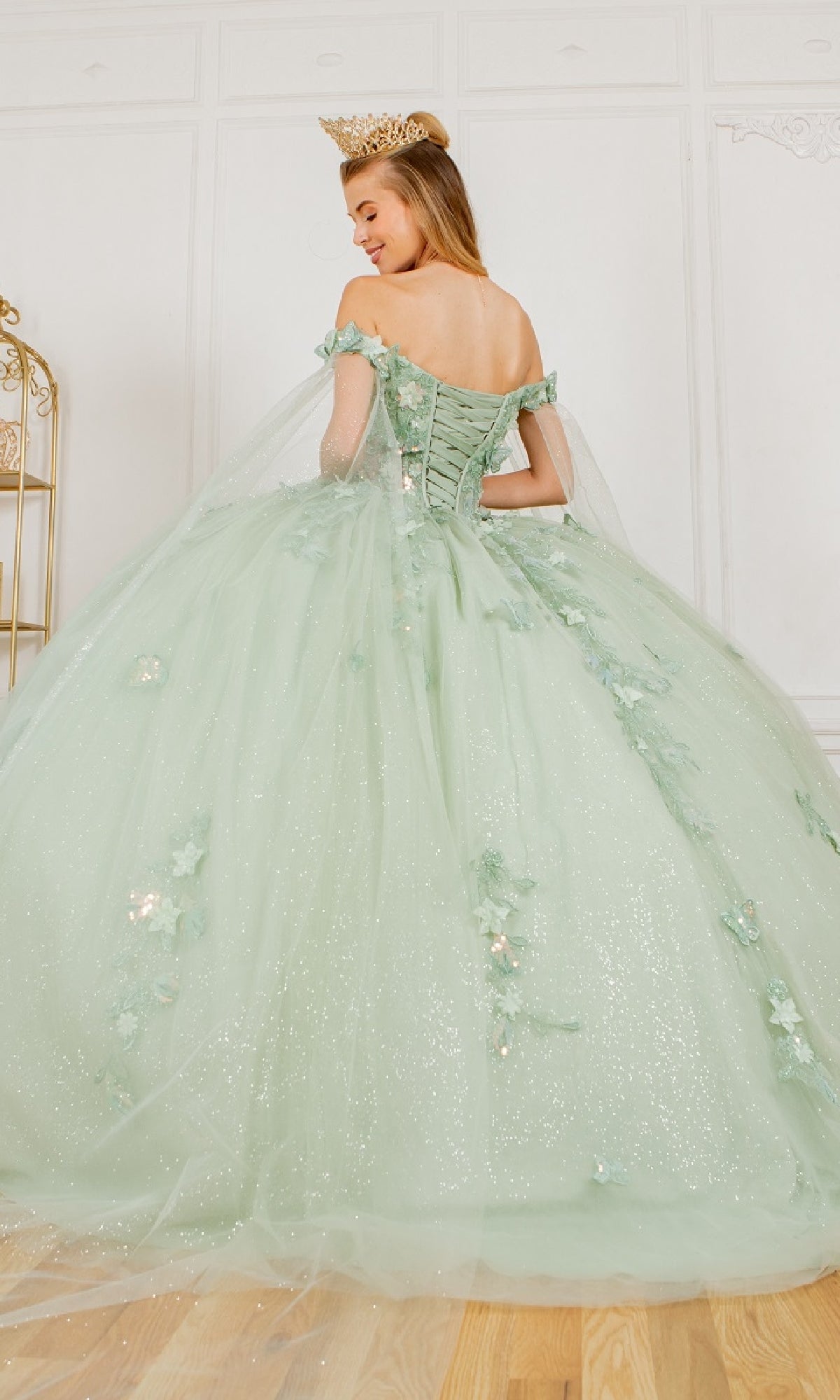 A woman in the Cinderella Couture 8075J Quinceanera Dress with off-the-shoulder 3D appliques and floral sparkles, wearing a gold crown, stands facing away in an elegant white room.